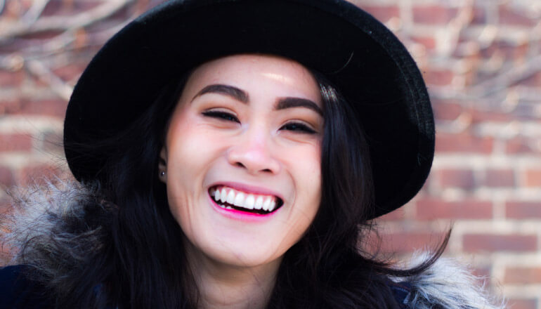 dark haired girl with black hat, coat with fur hood, and white teeth smiling