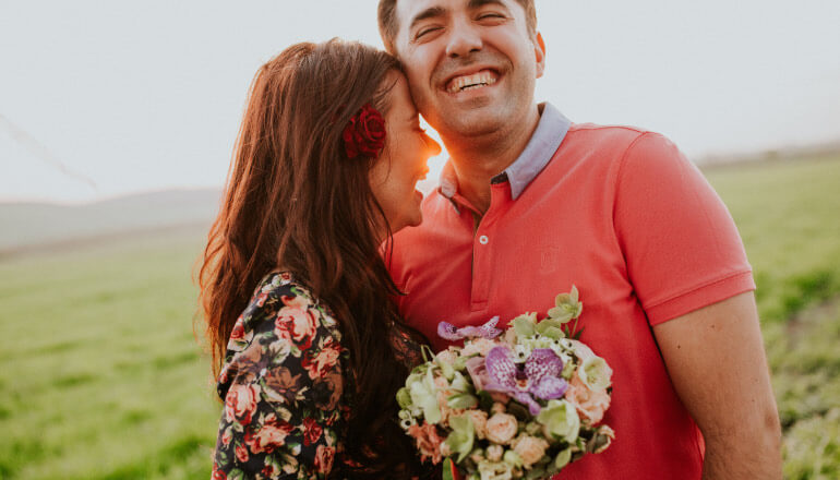 A dark-haired woman wearing a floral shirt holds a bouquet and embraces a man wearing a coral polo in a green field