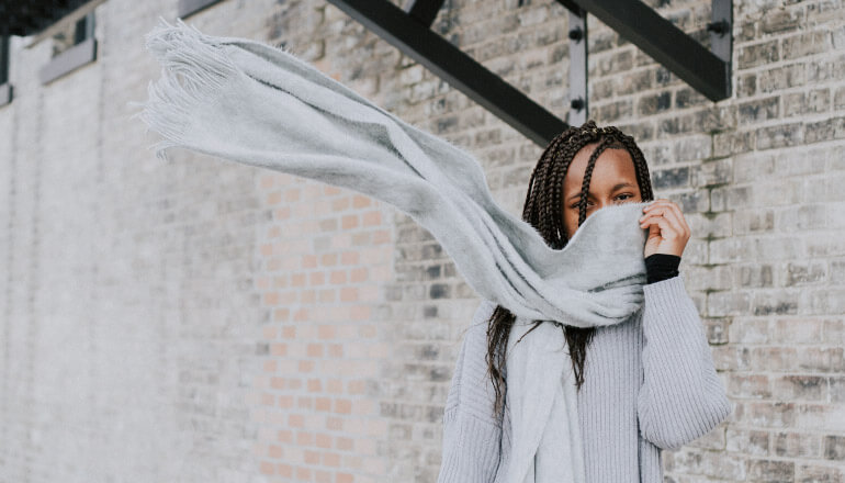 Dark-haired woman with multiple braids covers her mouth with a windblown light gray scarf in front of a gray brick wall