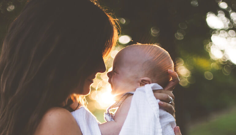 Closeup of a brunette mother holding her young baby in a white blanket as the sun sets