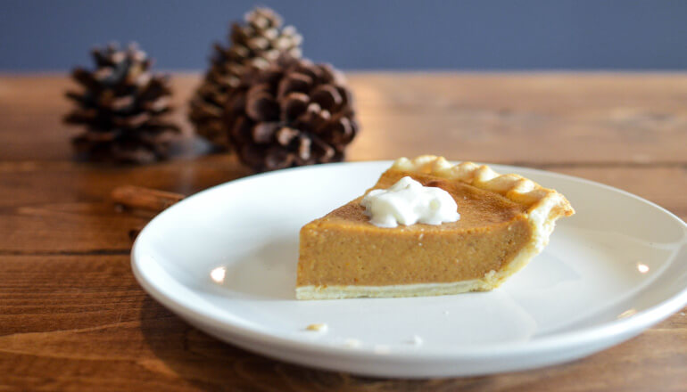 A piece of holiday pumpkin pie with a dollop of whipped cream on a white plate next to 3 pine cones on a wooden counter