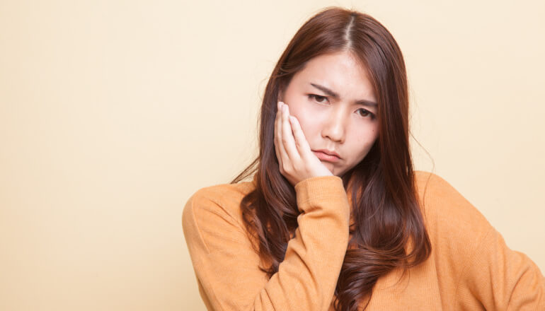 Brunette woman wearing a yellow sweater cringes in pain and touches her cheek due to oral pain from a sinus infection