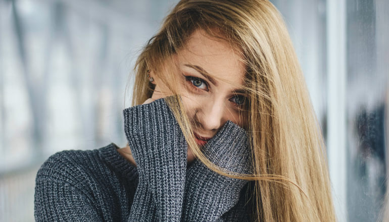 Blonde woman with sensitive teeth due to cold weather wears a gray sweater and covers her cheeks