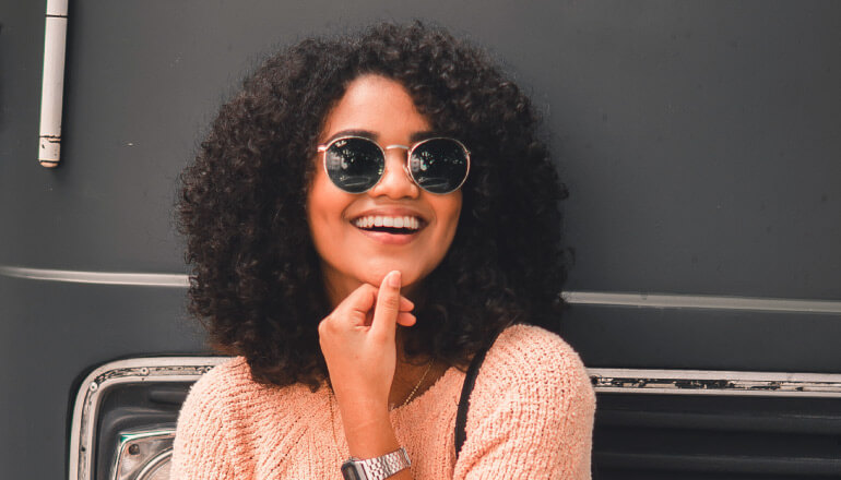 Curly-haired woman smiles with veneers while wearing an apricot sweater and sunglasses and standing against a black bus
