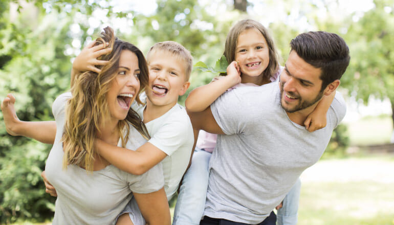 A mom and dad give piggy-back rides to their 2 kids while smiling and puling silly faces outside