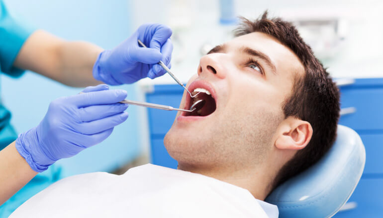 Brunette man getting his teeth cleaned at the dentist by a hygienist in blue gloves