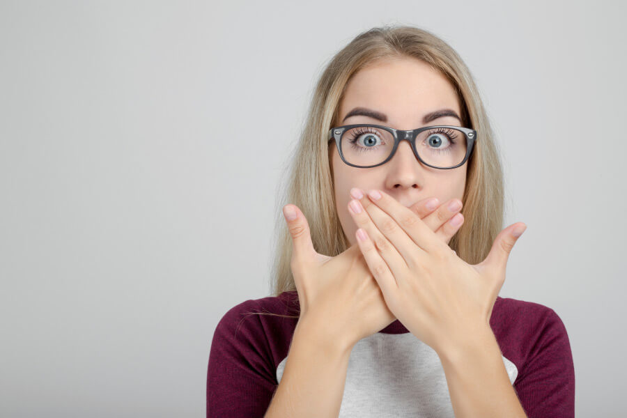 young woman wearing glasses covers her mouth with both hands to hide bad breath