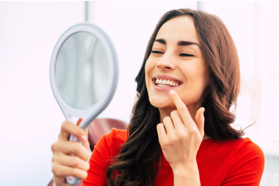 woman wearing a red dress looks in a handheld mirror to inspect the health of her gums