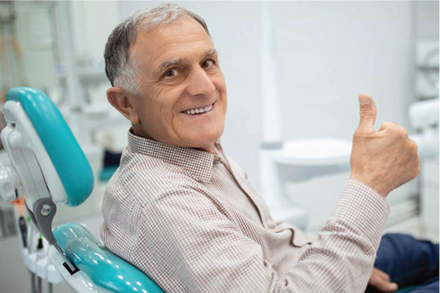 senior man sitting in the dentist chair gives a thumbs up after getting a dental bridge