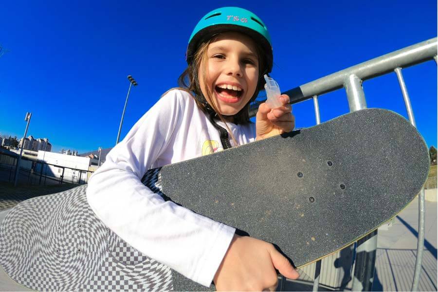 young girl wearing a helmet and holding a skateboard smiles and holds up her custom mouthguard
